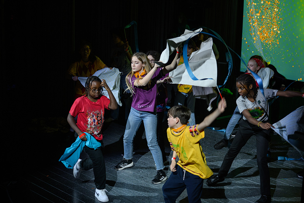 Children in multi-coloured costumes perform on a theatre stage.