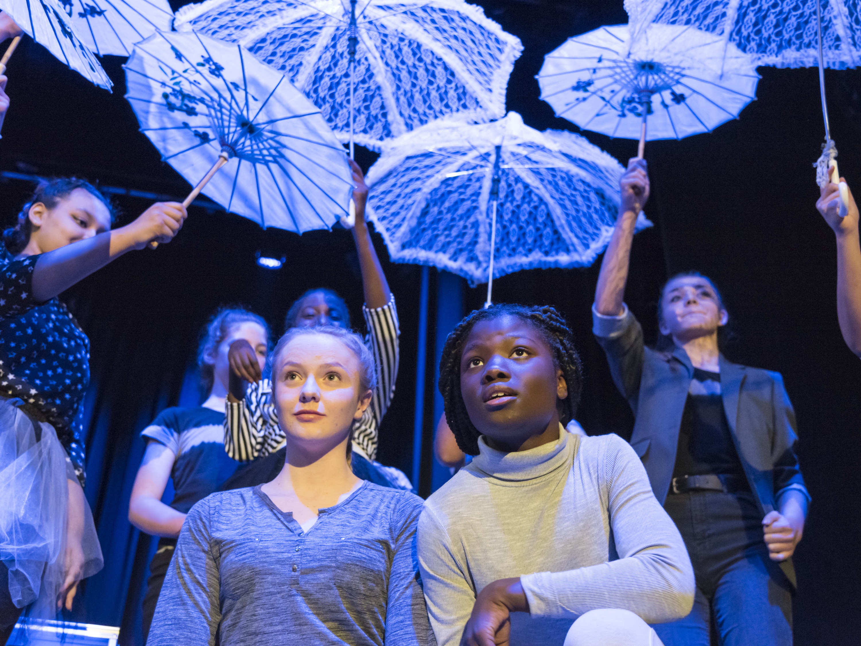 Teenagers on a theatre stage perform in front of brightly lit umbrellas.
