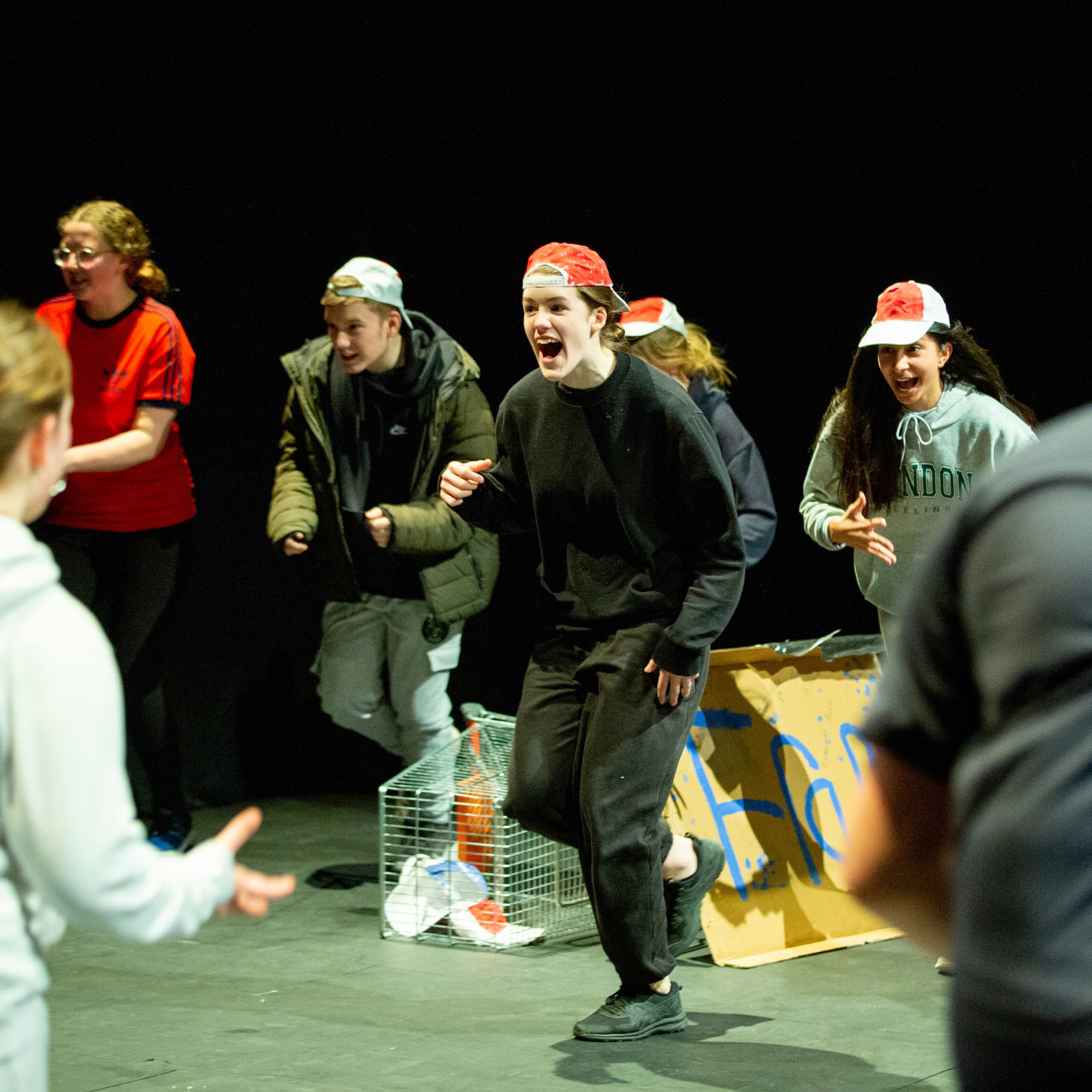 Smiling teenagers in costumes and backwards baseball caps run at each other on a theatre stage.
